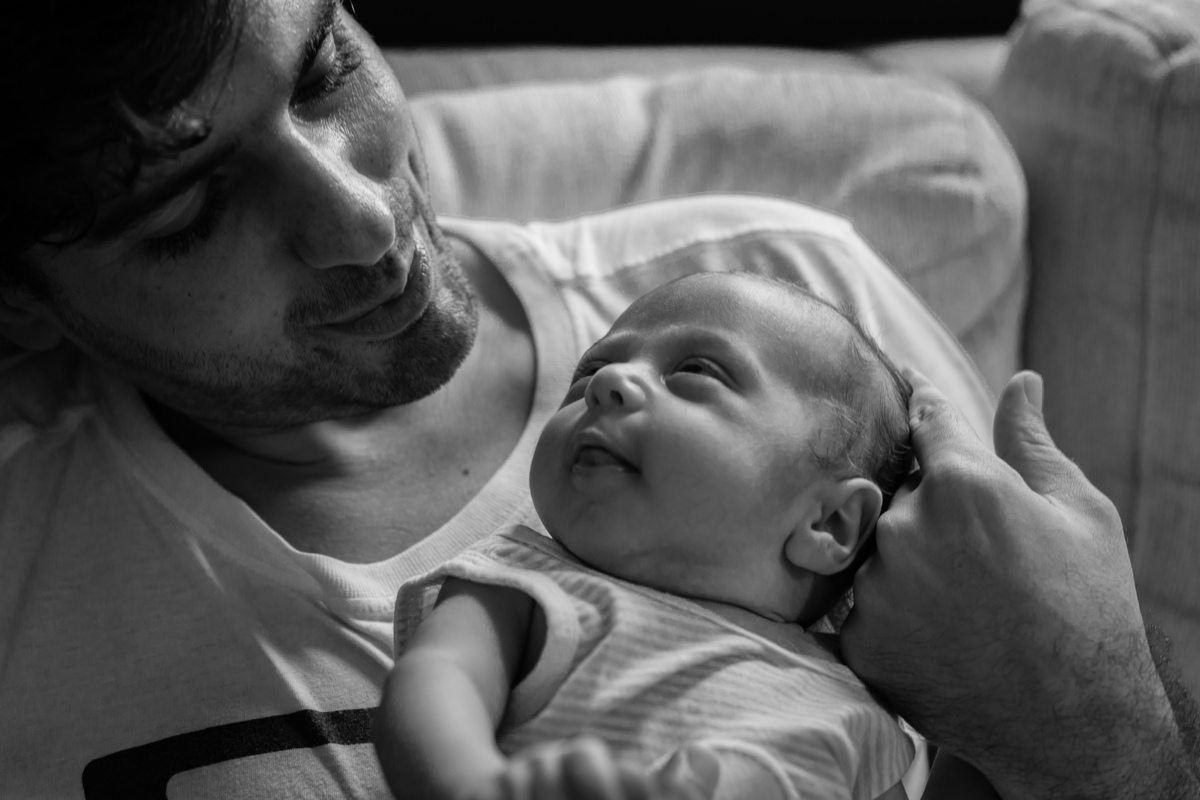 Papai sentado no sofá da sala com seu bebê, os dois se olham, o papai faz carinho em sua cabeça com muito amor