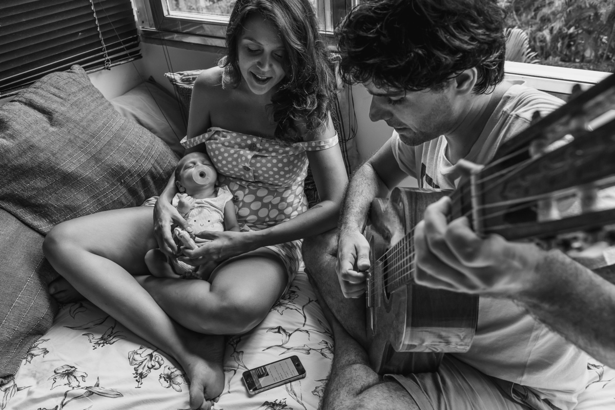Papai e mamãe fazem o bebê dormir, estão sentando em cima da cama, a mamãe embala o bebê enquanto o papai toca o violão e canta musicas de ninar acompanhando a letra pelo celular