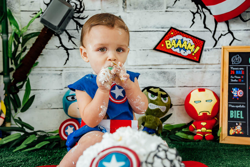 bebê fantasiado de capitão america no ensaio cake smash com o tema vingadores bebê com pirulito na mão lambusado de bolo do capitão america
