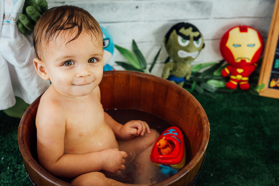 Bebê tomando banho em uma tina de madeira no ensaio cake smash, o bebê esta brincando com um barquinho vermelho no ensaio cake smash com o tema vingadores bebê