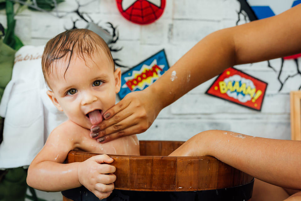 Bebê tomando banho em uma tina de madeira no ensaio cake smash com o tema vingadores bebê, o bebê esta com  a língua para fora 