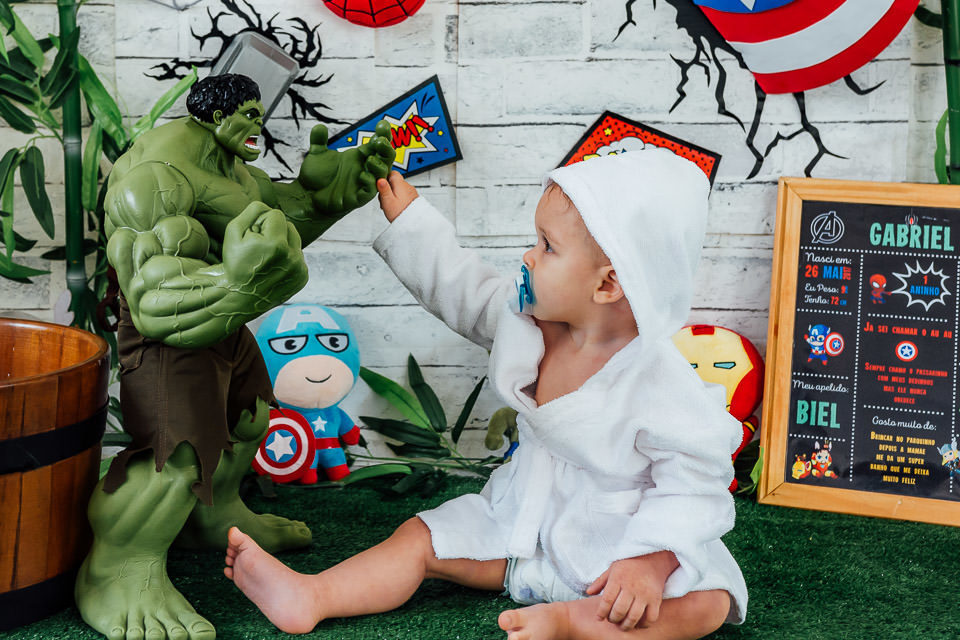 Bebê de roupão branco e com chupeta na boca, segurando a mão do hulk no ensaio cake smash com o tema vingadores bebê