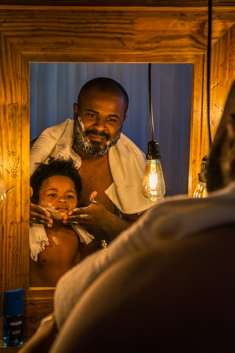 Pai e filho em frente ao espelho com moldura de madeira, o pai passa espuma de barbear no filho e ensina a fazer a barba.