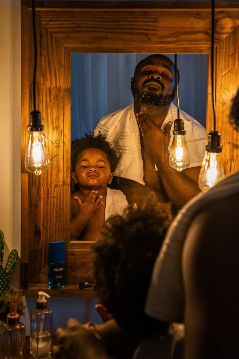 Pai e filho em frente ao espelho com moldura de madeira, o pai passa espuma de barbear no filho e ensina a fazer a barba.