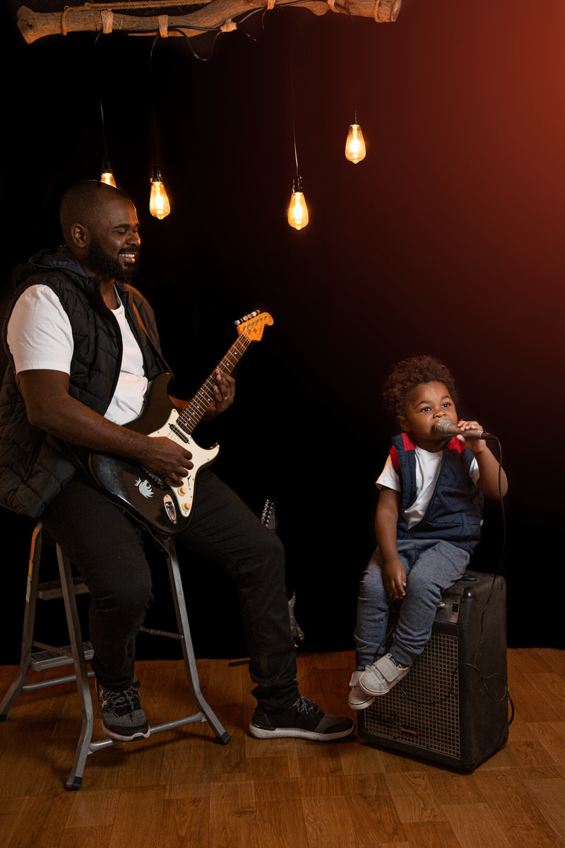 Pai e filho cantando, o pai tocando baixo e o filho no microfone sentado em uma caixa de som amplificadora.