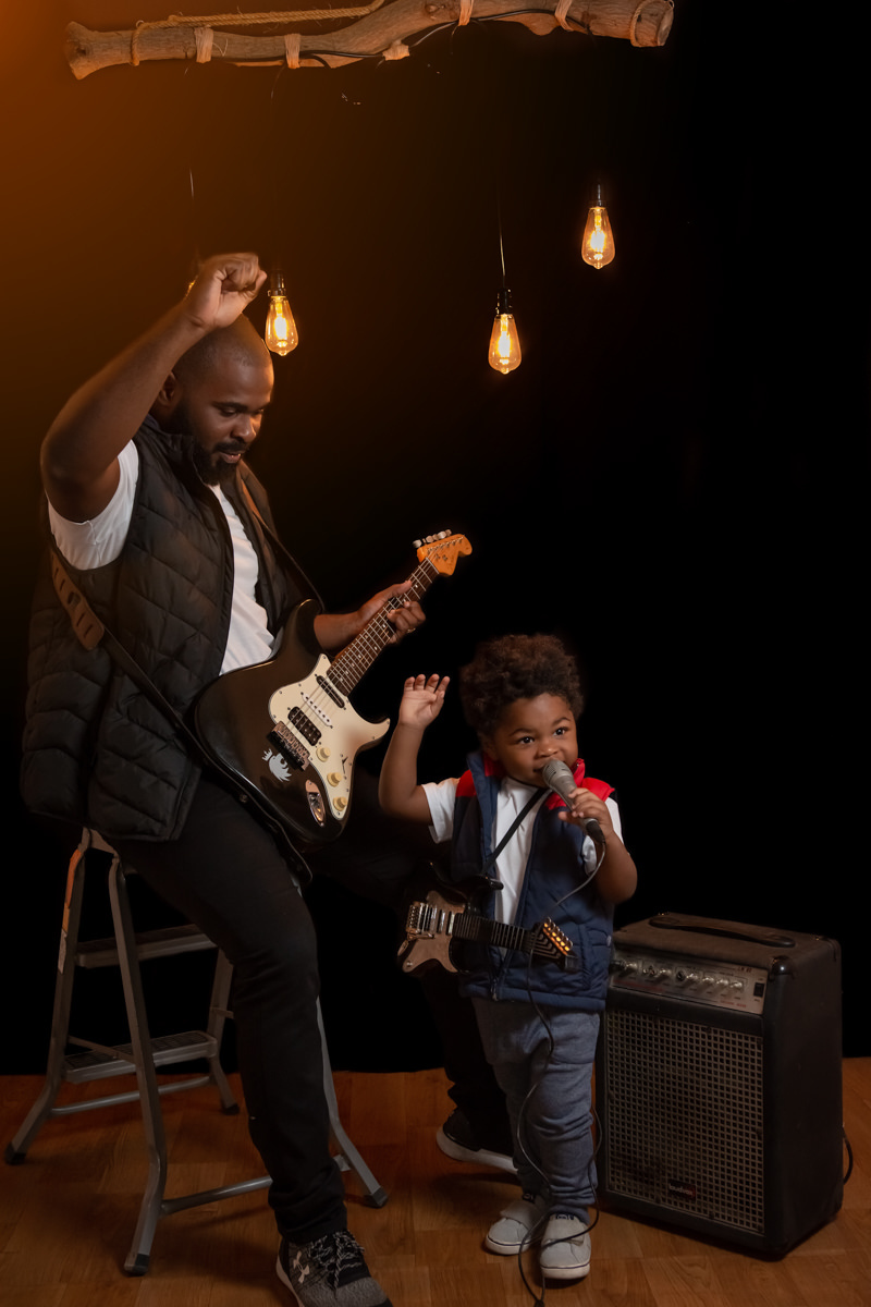 Pai e filho cantando, o pai tocando baixo e o filho no microfone. Os dois estão com as mão para o alto em agradecimento.