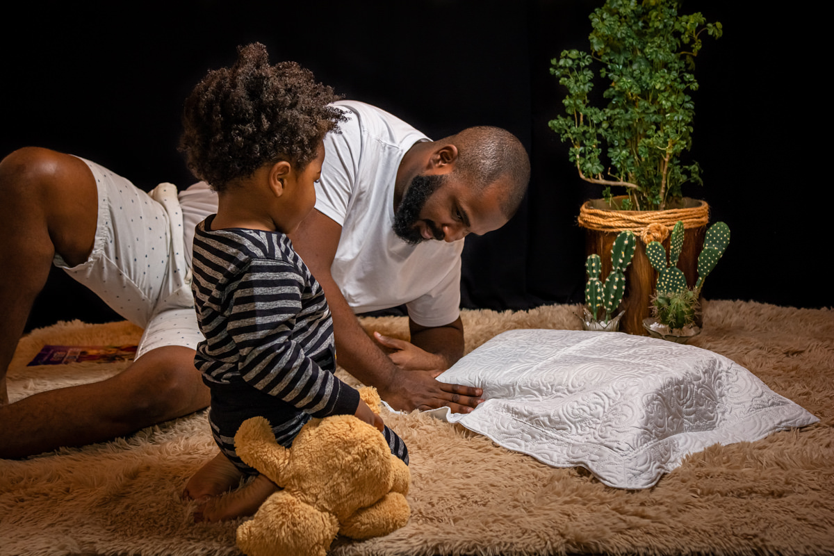 Pi e filho se preparam para dormir, o papai ajeita o travesseiro, o filho esta ajoelhado segurando o urso de pelúcia marrom e veste pijama listrado.