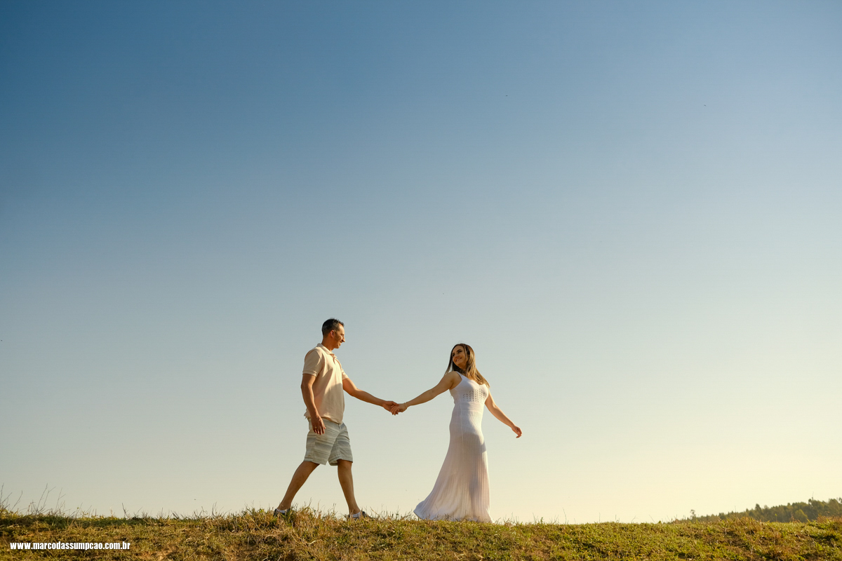 casal caminhando de maos dadas se olhando em cima de morro com ceu azul