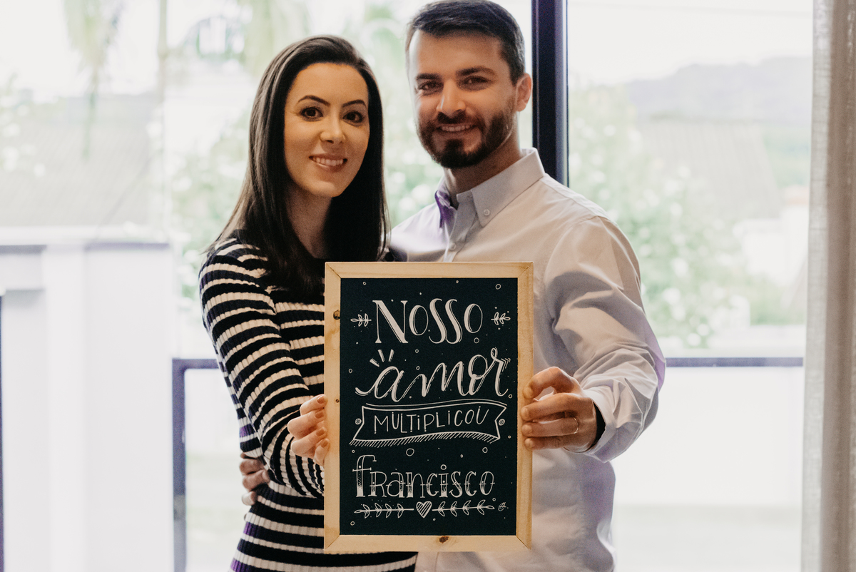 casal segurando a plaquinha com os dizeres nosso amor multiplicou, ela usa vestido listrado e ele camisa lisa, em frente a janela de vidro.