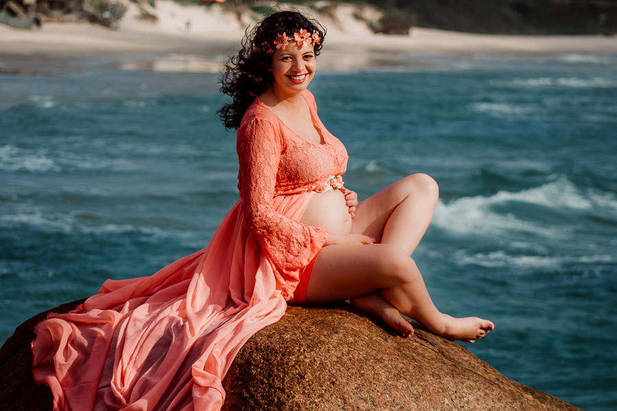 gravida sorrindo sentada sobre a pedra com mar ao fundo
