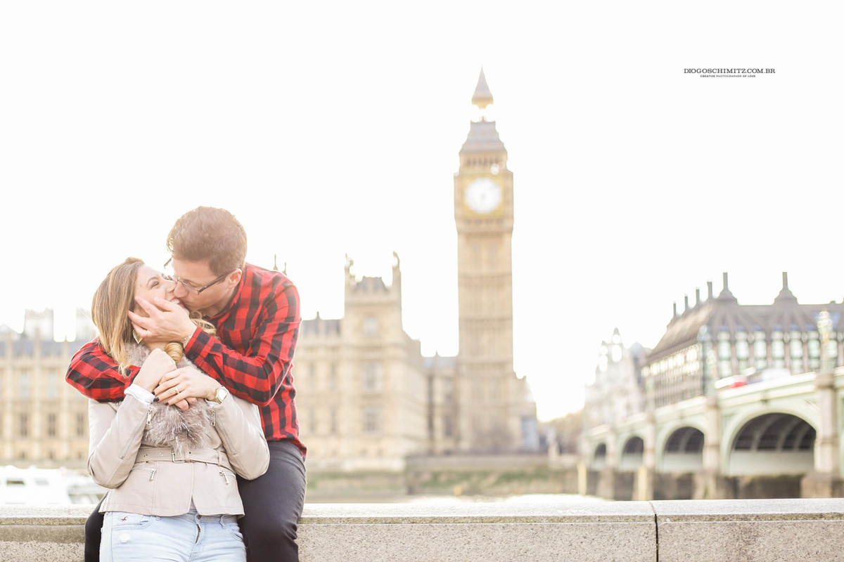 Homem sentando em uma mureta beijando a mulher, ao fundo da foto se avista o Palácio de Westminster e a Torre do Big Ben.