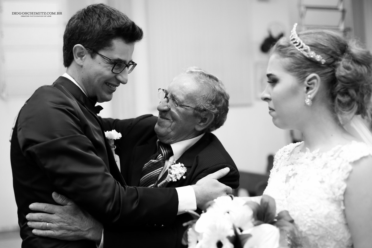 Noivo e o pai da noiva sorrindo enquanto a noiva os observa.