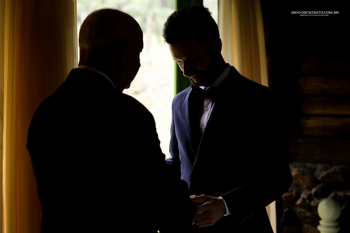 Noivo sorrindo ao ver o seu pai arrumando seu terno para o casamento no campo.