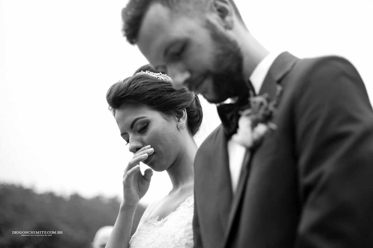 Noiva segura o choro com a mão, noivo ao seu lado reza.