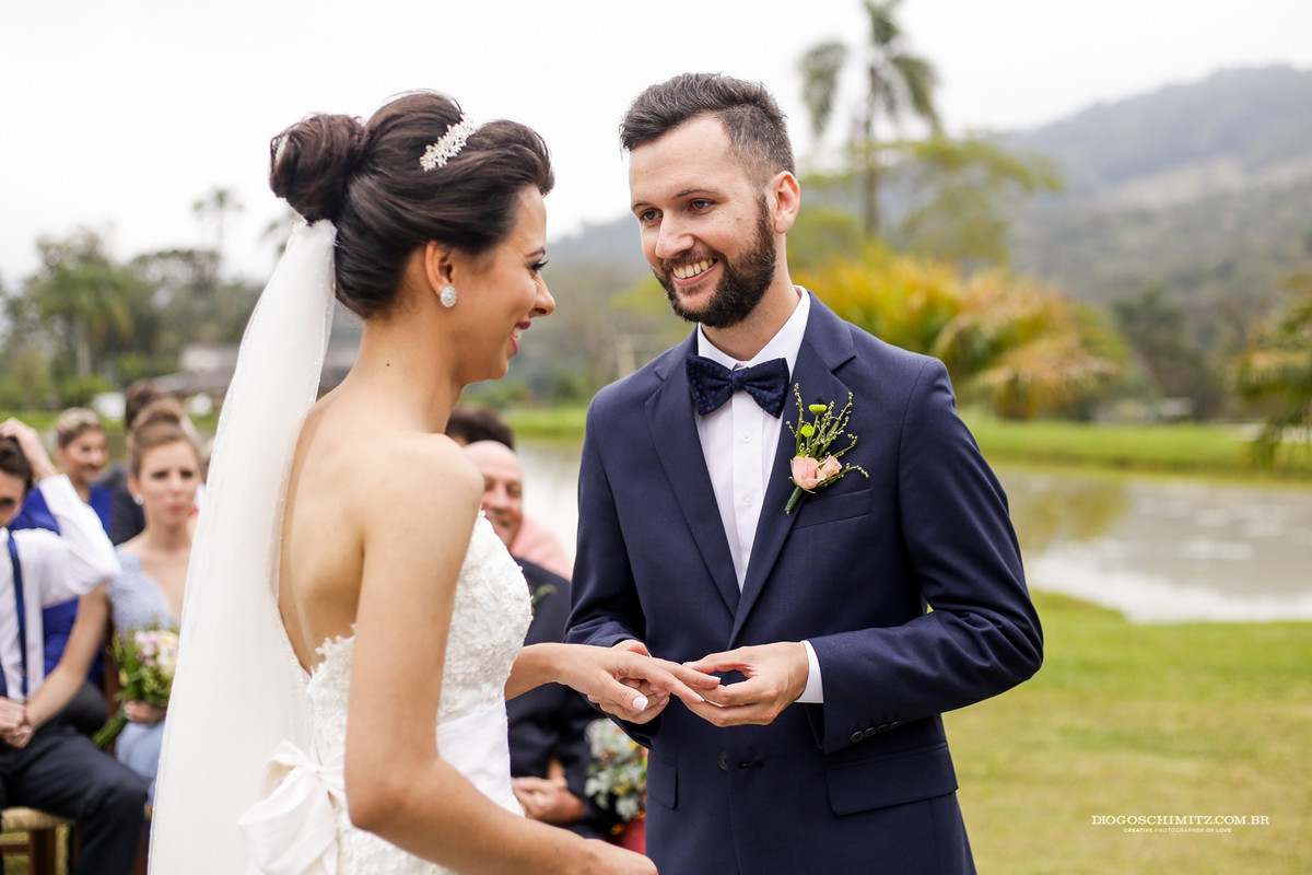 Noivo sorri para a noivo no momento que coloca a aliança no dedo da noiva.