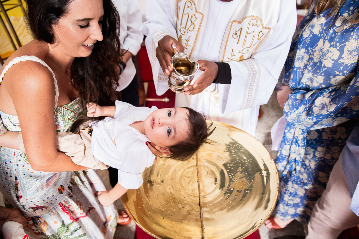 familia participando do batismo madrinha segurando criança enquanto o padre joga agua