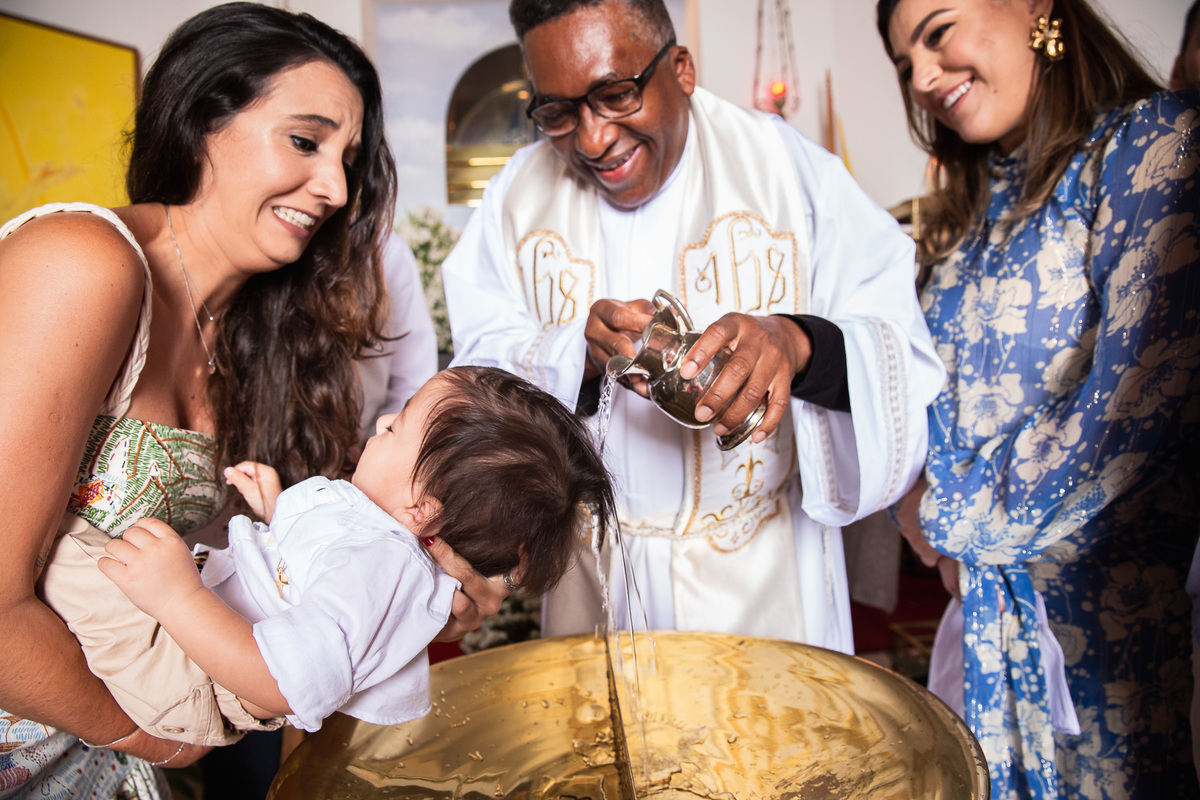 familia participando do batismo madrinha segurando criança enquanto o padre joga agua