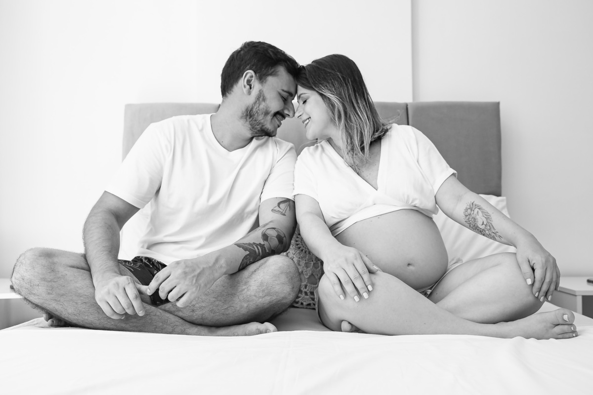 casal com 30 semanas de gestação posando testa com testa pra foto em cima da cama de casal