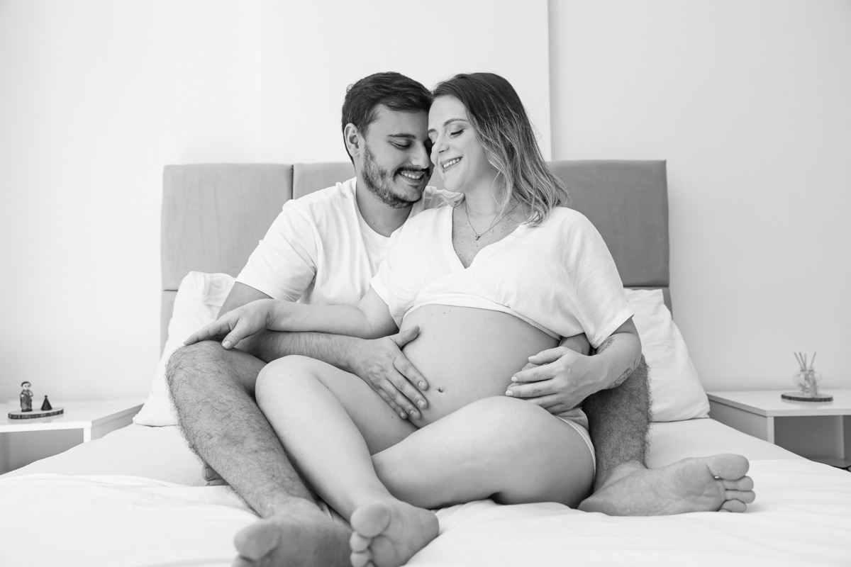 casal com 30 semanas de gestação posando pra foto testa com testa e com a mão na barriga em cima da cama de casal
