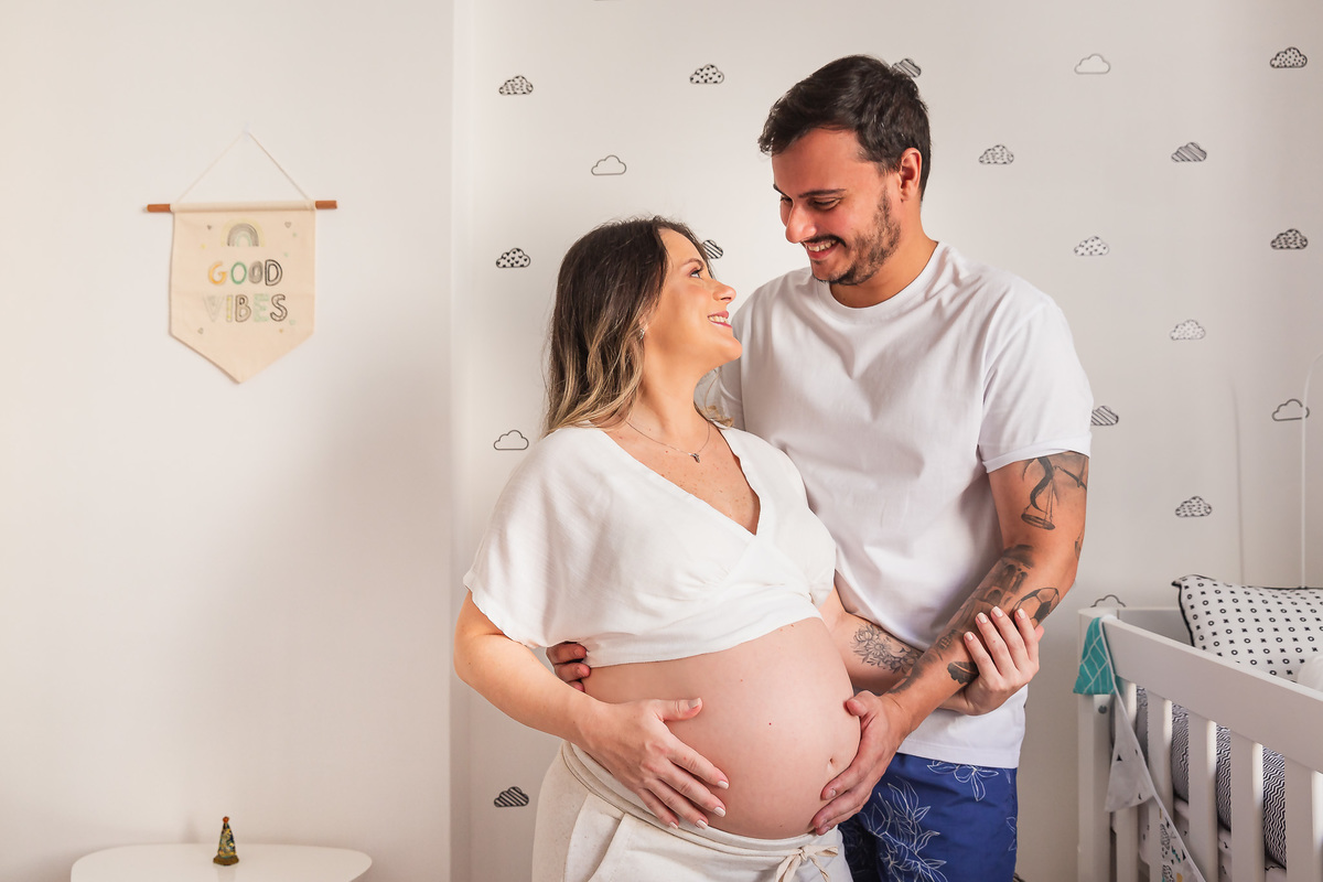 casal dentro do quarto do bebe com a mão na barriga da mãe posando pra foto