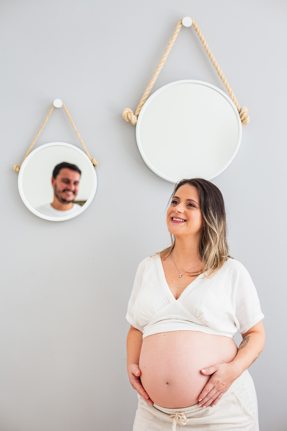 mãe com 30 semanas de gestação posando pra foto com a mão na barriga enquanto olha para o marido através do espelho redondo da sala