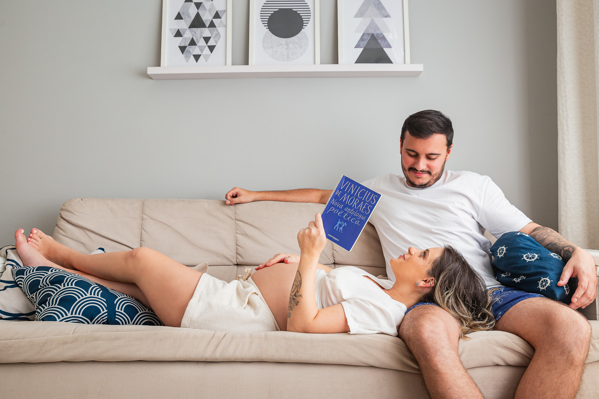 casal com 30 semanas de gestação posando pra foto ele sentado no sofa e ela deitada com a cabeça no colo dele lendo um livro de poemas do vinicius de moraes