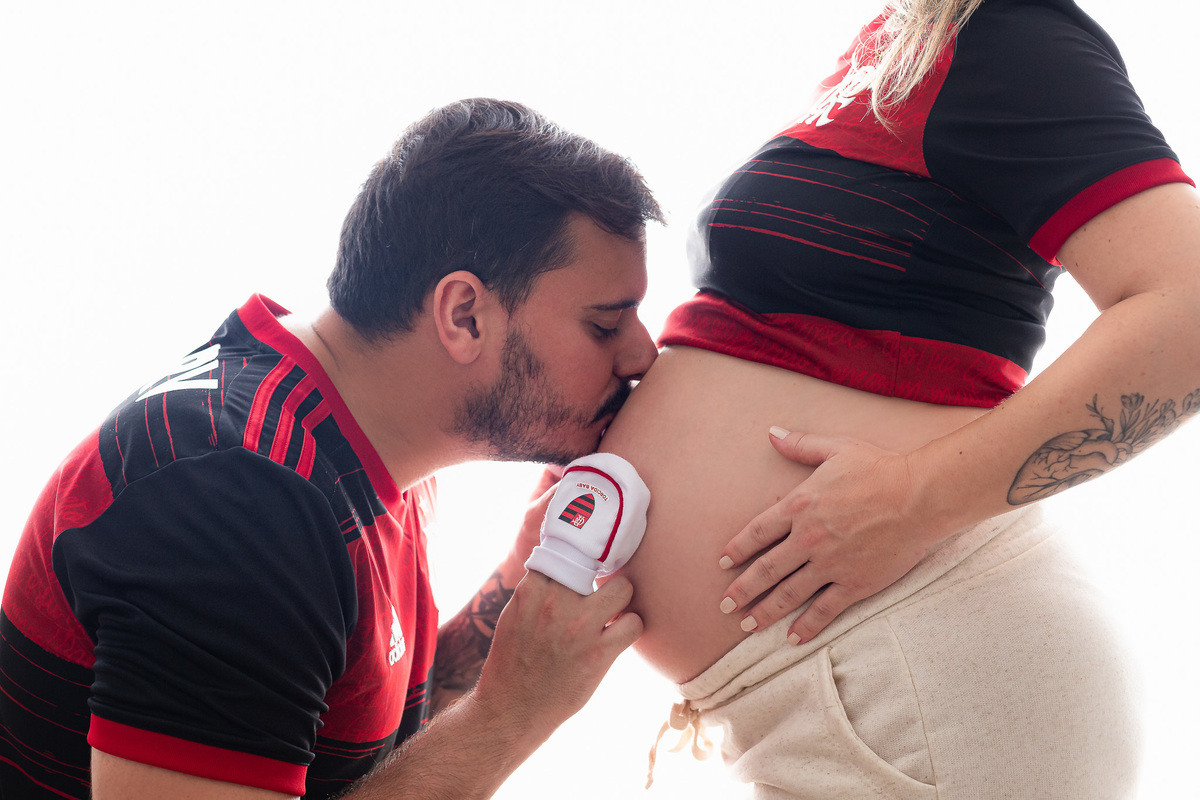 casal flamenguista com 30 semanas de gestação posando pra foto com um sapatinho do flamengo na barriga e beijando a barriga