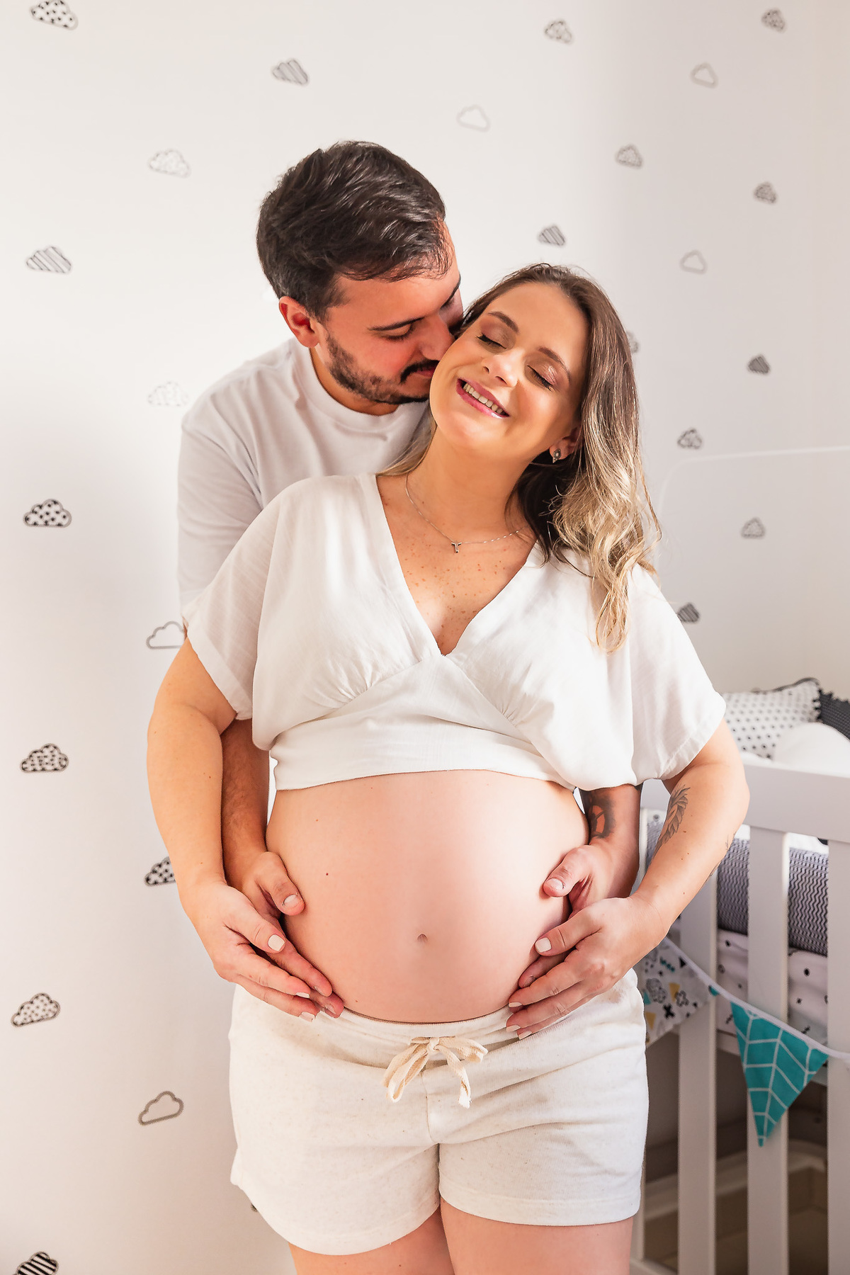 casal dentro do quarto do bebe com a mão na barriga da mãe posando pra foto