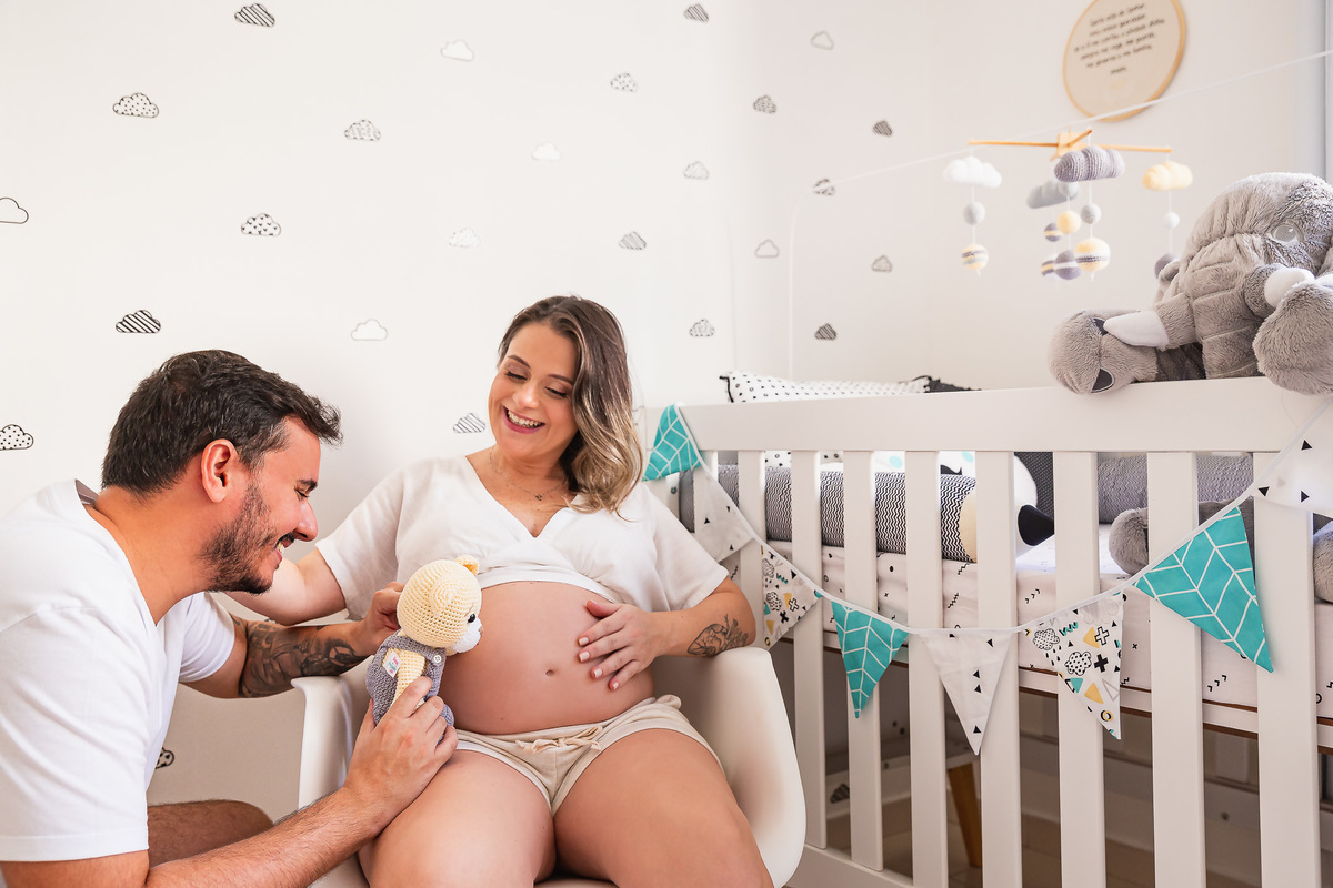 casal dentro do quarto do bebe pai e mae brincando com ursinho de pelucia na barriga
