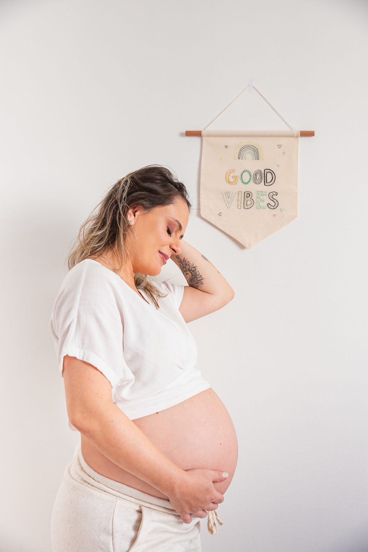 mãe com 30 semanas de gestação posando pra foto com a mão na barriga e com uma flamula de good vibes ao fundo