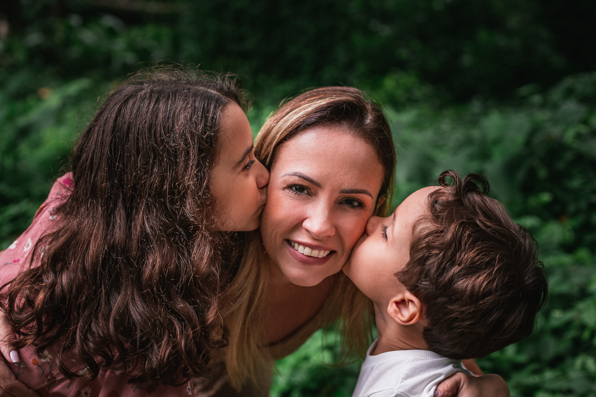 Ensaio fotografico de familia no parque lage mae recebendo beijo dos filhos com folhas verdes de fundo