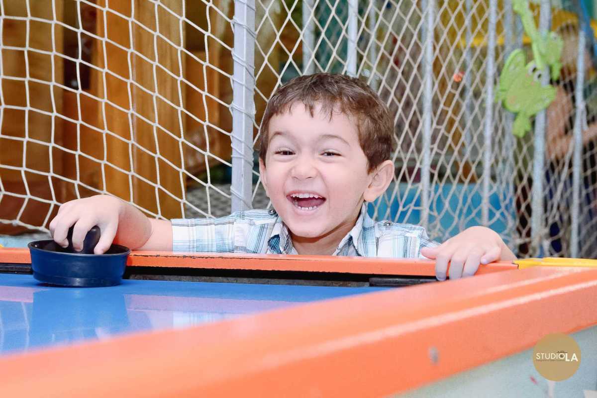 Menino jogando air hockey com sorriso durante sua festa infantil no Buffet Azul do Mar em São Paulo. Fotografia alegre feita pelo Studio LA.