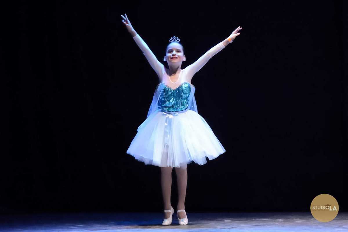 Fotografia de apresentação de dança infantil, com bailarina mirim em figurino branco realizando movimento clássico no palco. Registro artístico feito pelo Studio LA durante espetáculo de dança e teatro.