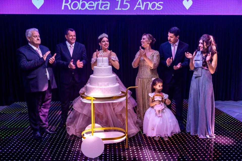 Hora do Parabéns na festa de 15 anos da debutante Roberta, com a família reunida em volta do bolo. Fotografia de festa de 15 anos em São Paulo.