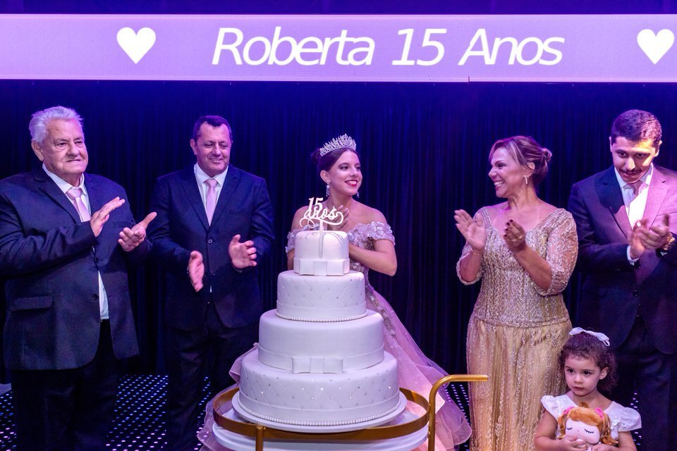 Hora do Parabéns na festa de 15 anos da debutante Roberta, com a família reunida em volta do bolo. Fotografia de festa de 15 anos em São Paulo.