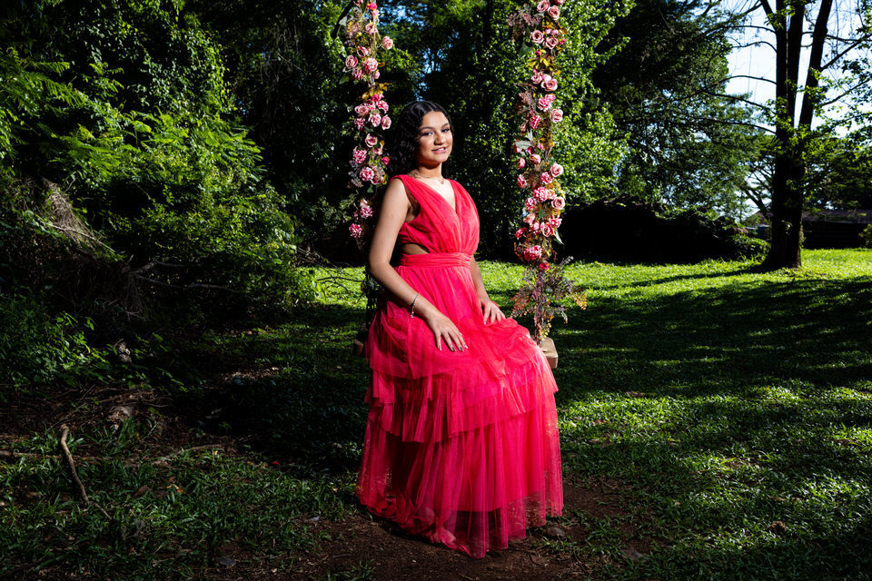 Fotógrafo de debutante em Holambra registra jovem com vestido pink sentada em um balanço decorado com flores, em meio à natureza. Macena Flores 