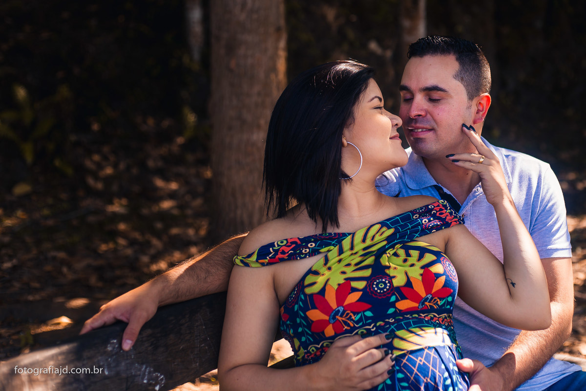 gestante e marido sentados em um banco de madeira. A grávida está com as pontas dos dedos no rosto do marido e os dois estão quase se beijando.