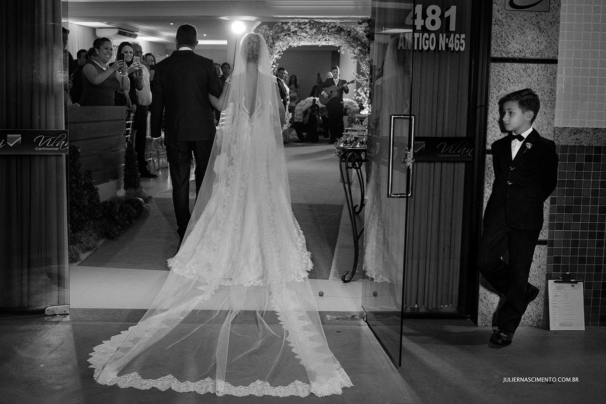 fotografia preto e branco da entrada da noiva no cerimonial Vilani em Vitória-ES, e pajem olhando para o véu da noiva.
