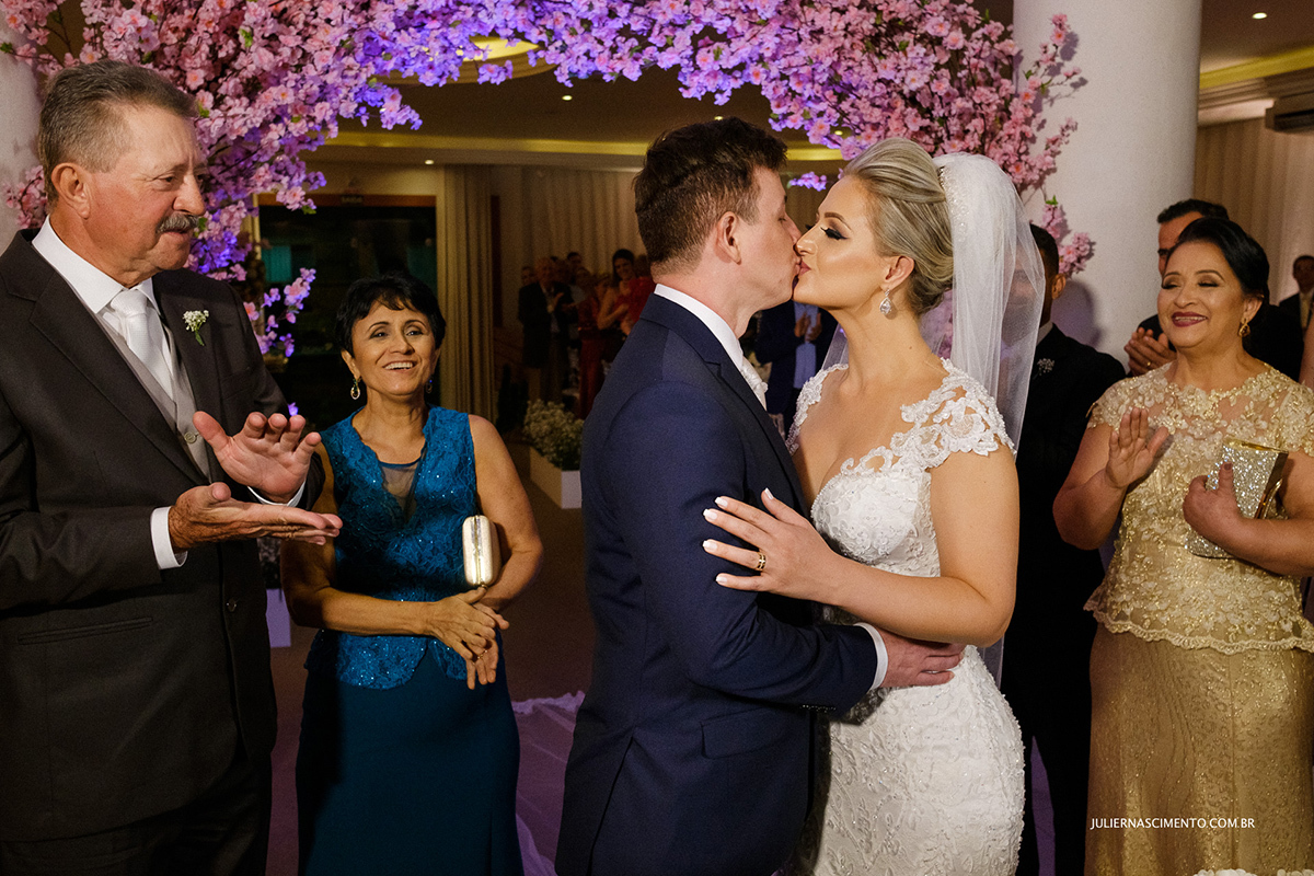 Foto do beijo dos noivos no dia de casamento e pais aplaudindo.
