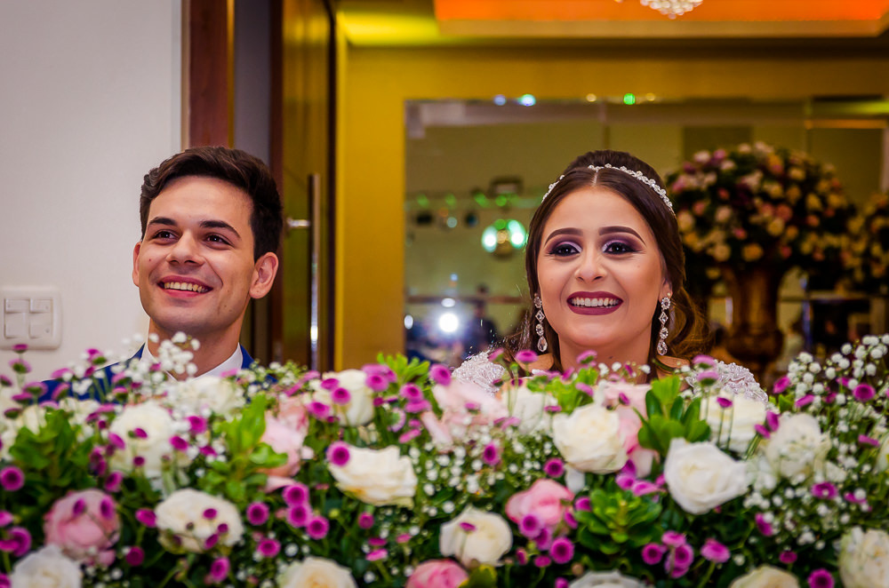 Fotografia da recepção ou festa de casamento dos noivos sentados e sorrindo e flores brancas e coloridas como decoração da mesa no espaço de eventos em Maringá PR
