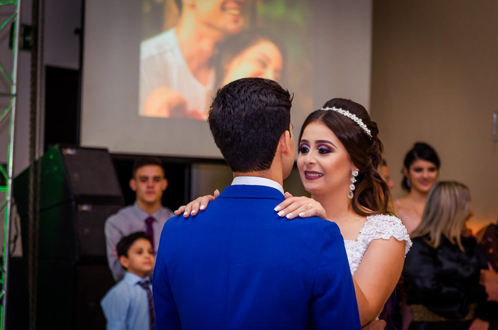 Fotografia da recepção ou festa de casamento da dança dos noivos com os convidados ao fundo. Ele de terno azul e a noiva de vestido de noiva branco e longo com rendas e uma tiara no cabelo no espaço de eventos em Maringá PR