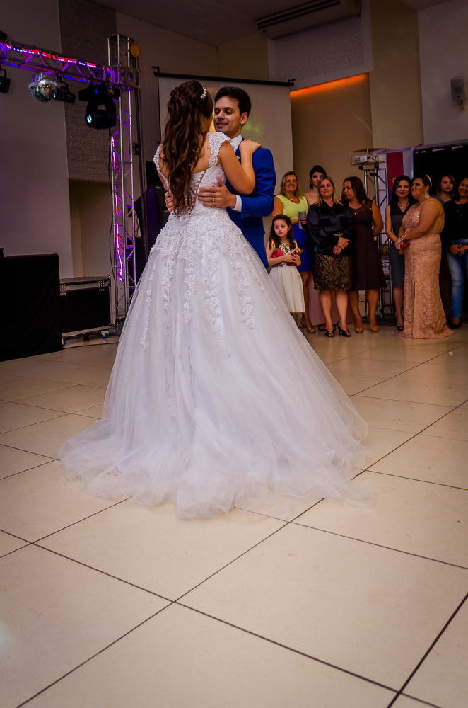 Fotografia da recepção ou festa de casamento da dança dos noivos com os convidados ao fundo. Ele de terno azul e a noiva de vestido de noiva branco e longo com rendas e uma tiara no cabelo no espaço de eventos em Maringá PR