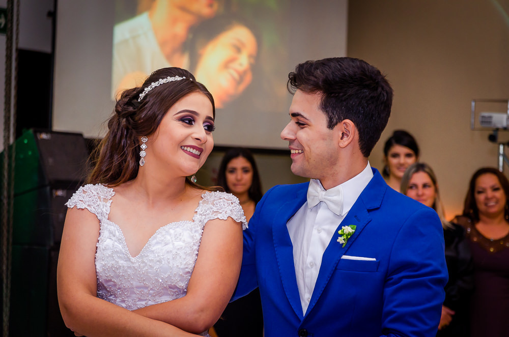Fotografia da recepção ou festa de casamento da dança dos noivos com os convidados ao fundo. Ele de terno azul e a noiva de vestido de noiva branco e longo com rendas e uma tiara no cabelo no espaço de eventos em Maringá PR