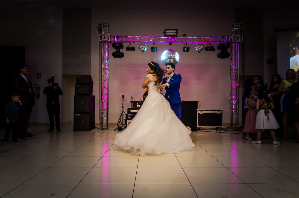 Fotografia da recepção ou festa de casamento da dança dos noivos com os convidados ao fundo. Ele de terno azul e a noiva de vestido de noiva branco e longo com rendas e uma tiara no cabelo no espaço de eventos em Maringá PR