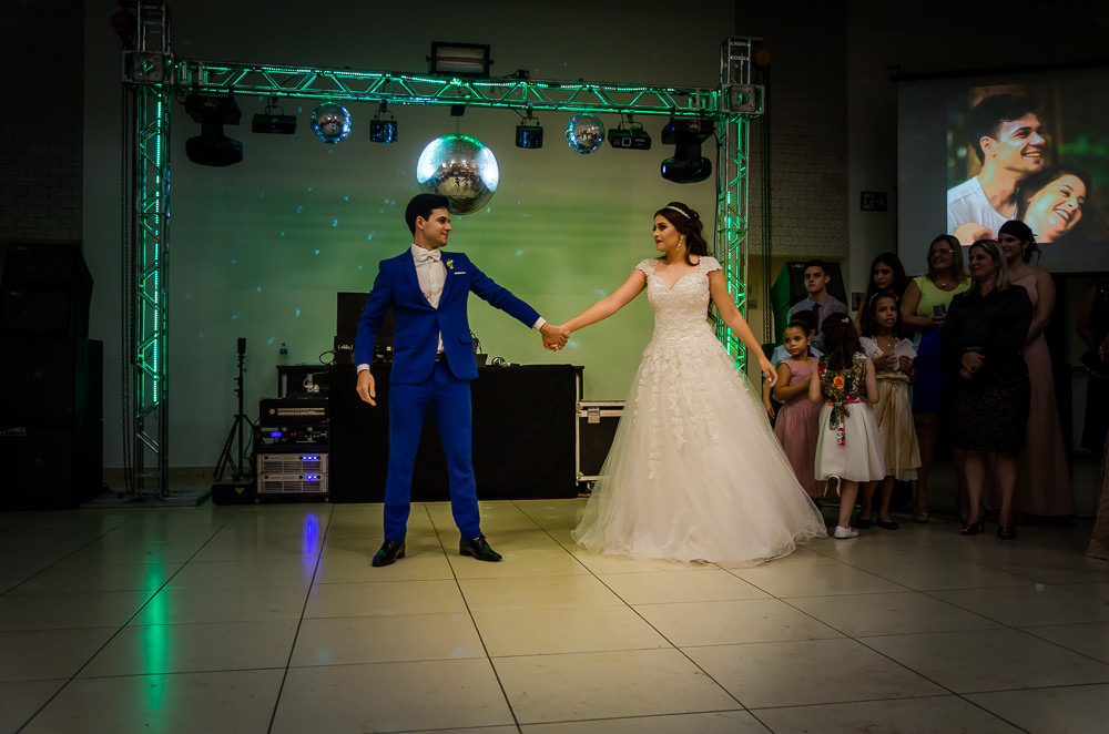 Fotografia da recepção ou festa de casamento da dança dos noivos com os convidados ao fundo. Ele de terno azul e a noiva de vestido de noiva branco e longo com rendas e uma tiara no cabelo no espaço de eventos em Maringá PR