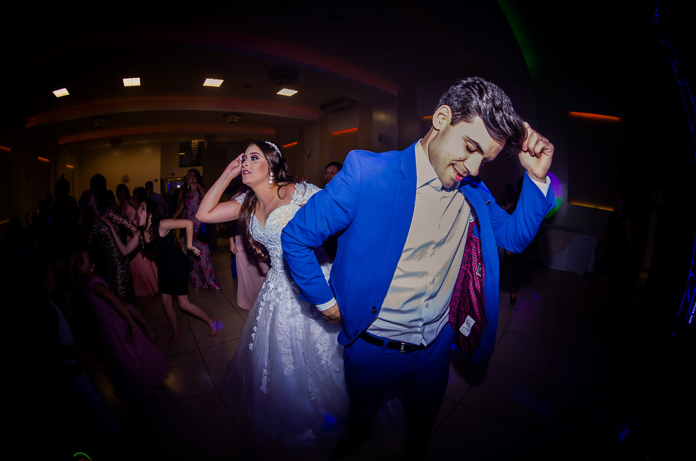 Fotografia da recepção ou festa de casamento da dança dos noivos com os convidados ao fundo. Ele de terno azul e a noiva de vestido de noiva branco e longo com rendas e uma tiara no cabelo no espaço de eventos em Maringá PR