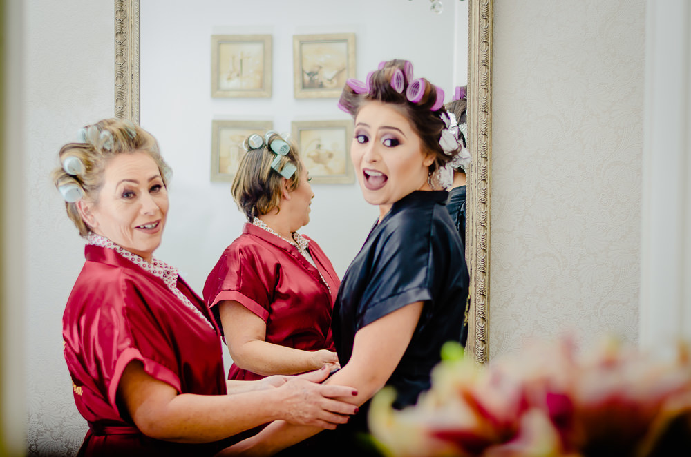Fotografia na noiva no making of no dia do casamento no salão de beleza no dia da noiva abraçando sua ma~e e ambas com olhar surpreso quando chega o buquê ou buquet da noiva em Maringá PR