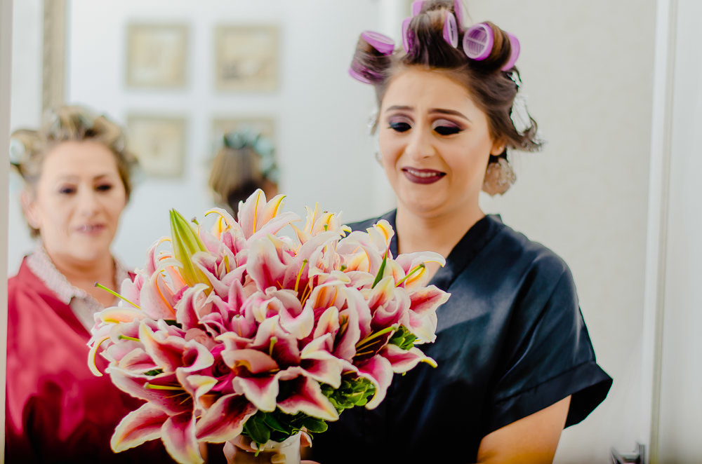 Fotografia na noiva no making of no dia do casamento no salão de beleza no dia da noiva com sua mãe ao fundo. Ela olha admirada para seu buquê ou buquet de casamento com lírios em Maringá PR