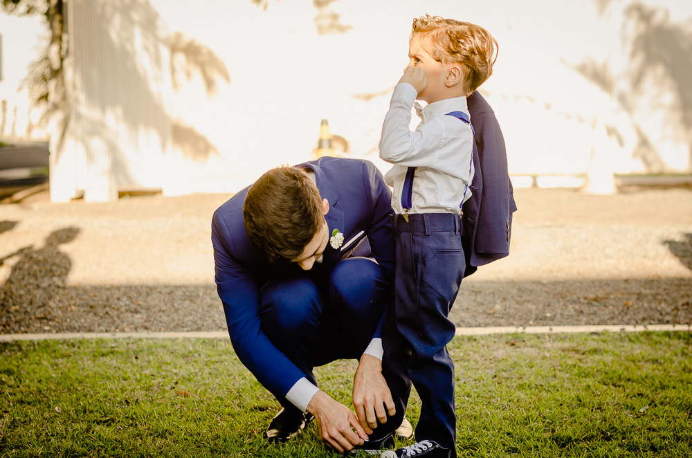 Fotografia do noivo usando terno azul amarrando o cadardo do sapado do pajem numa cerimônida de casamento durando o dia, com sol e sob um gramado verde no espaço de eventos em Maringá PR