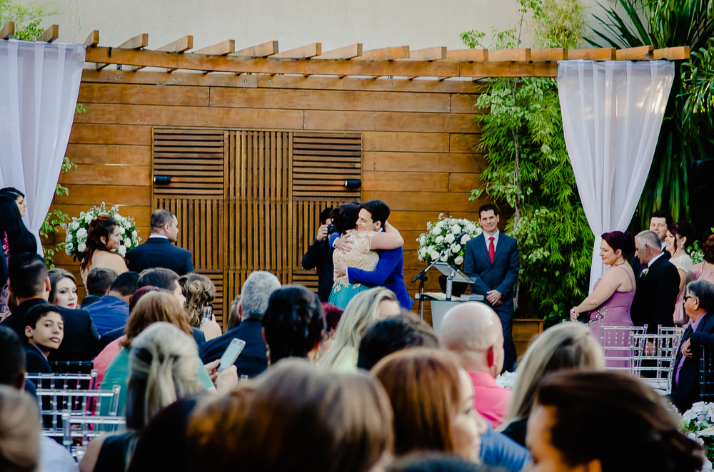 Fotografia do noivo no dia da cerimônia de casamento vestindo terno azul abraçando sua mãe emocionados, vida de tráz com todos os convidados. Cerimônia durante o dia a céu aberto e ensolarado no espaço de eventos em Maringá PR